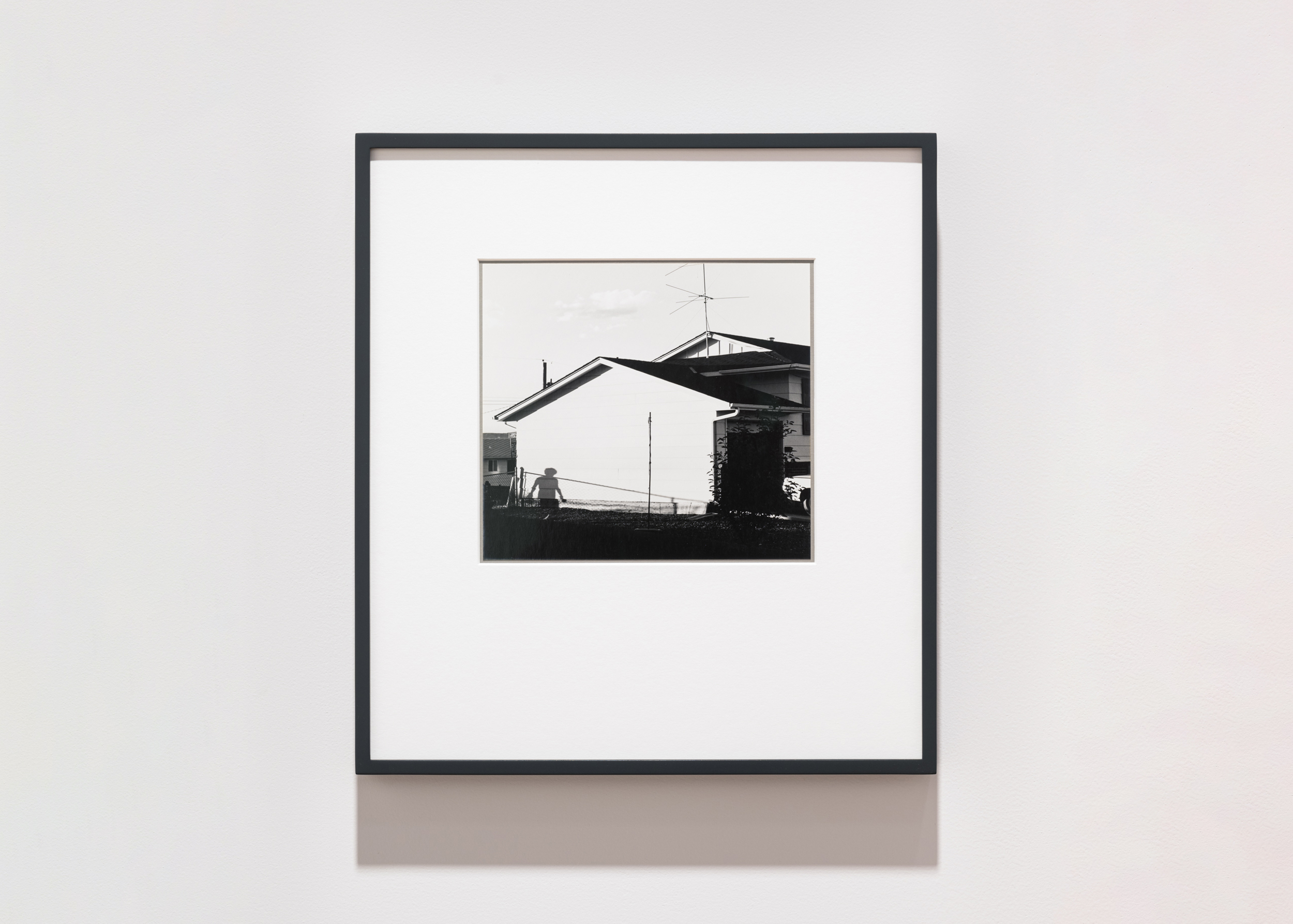 Color image of a black and white photograph of the exterior of a home with the shadow of a figure walking framed in black on a white wall