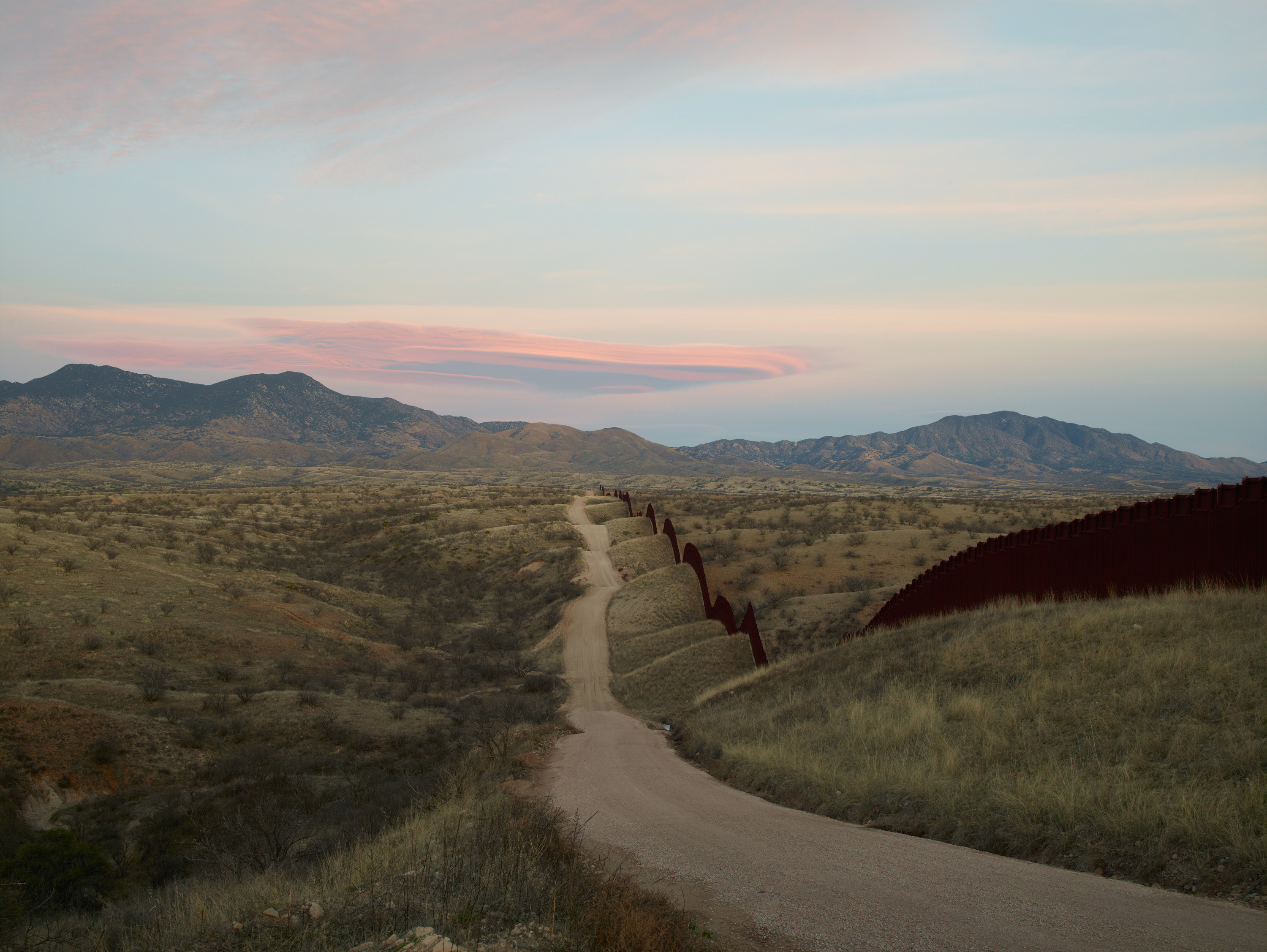 Color photograph of the US/Mexico border with a mountain range in the distance with pinkish blue skies