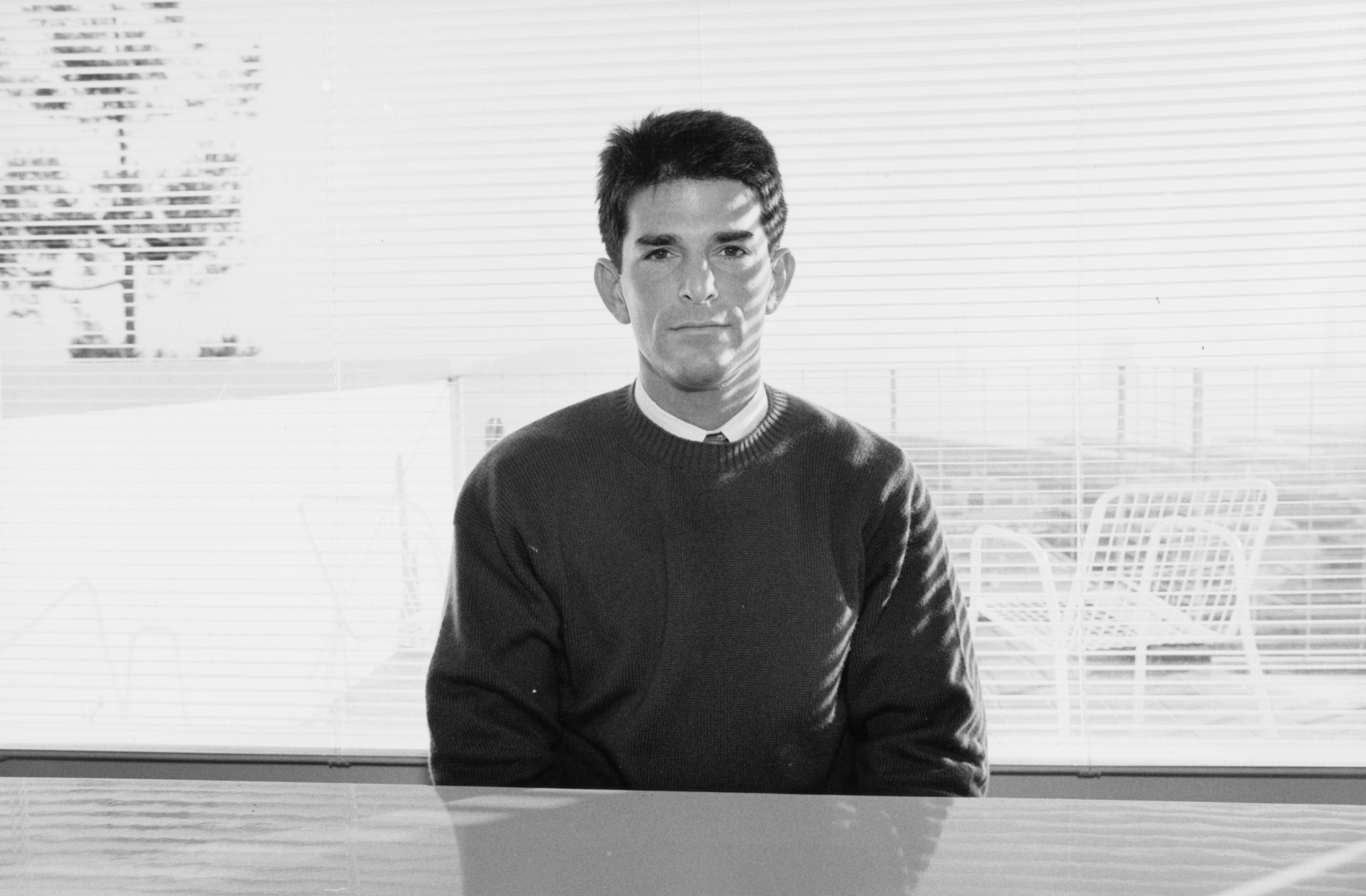 Black and white photograph of a seated figure behind a desk looking at the viewer