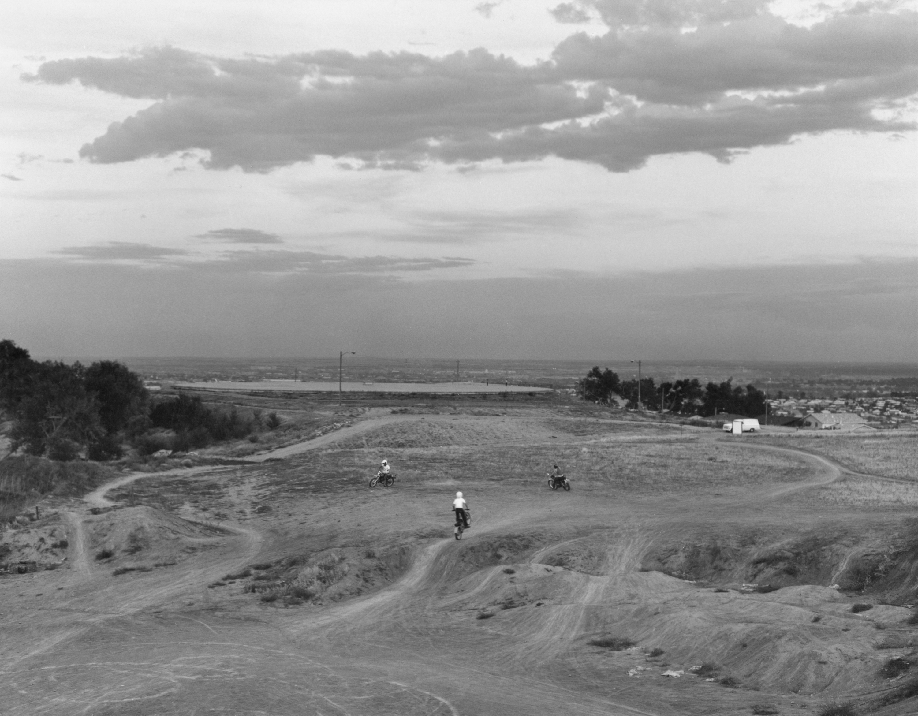 Black and white photograph of three figures on motorbikes riding around a dirt field