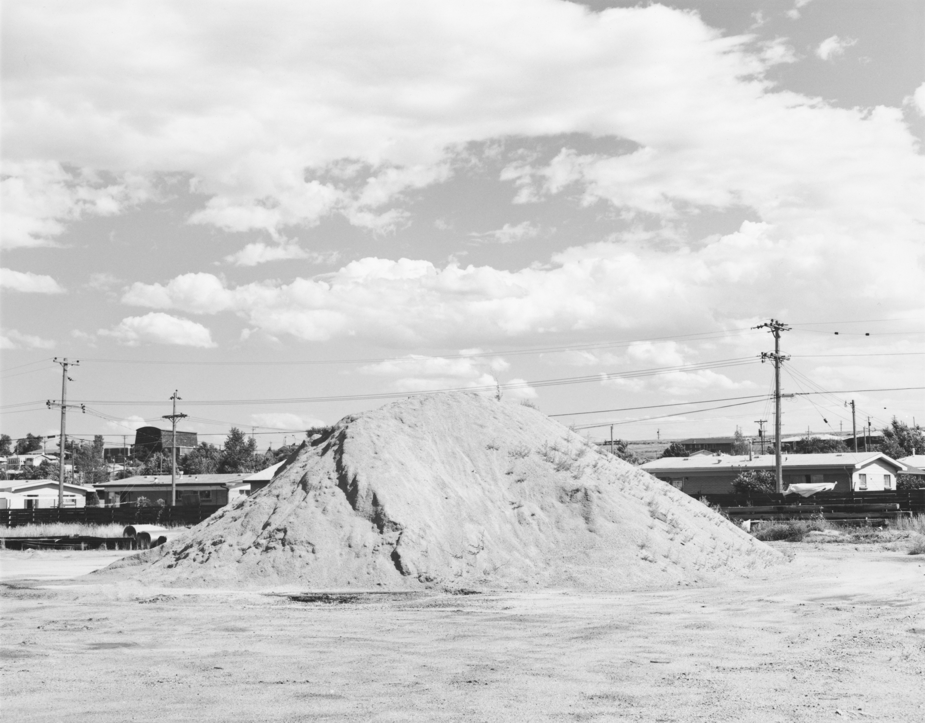 Black and white photograph of a mound of dirt in the middle of an abandon lot amongst residential homes