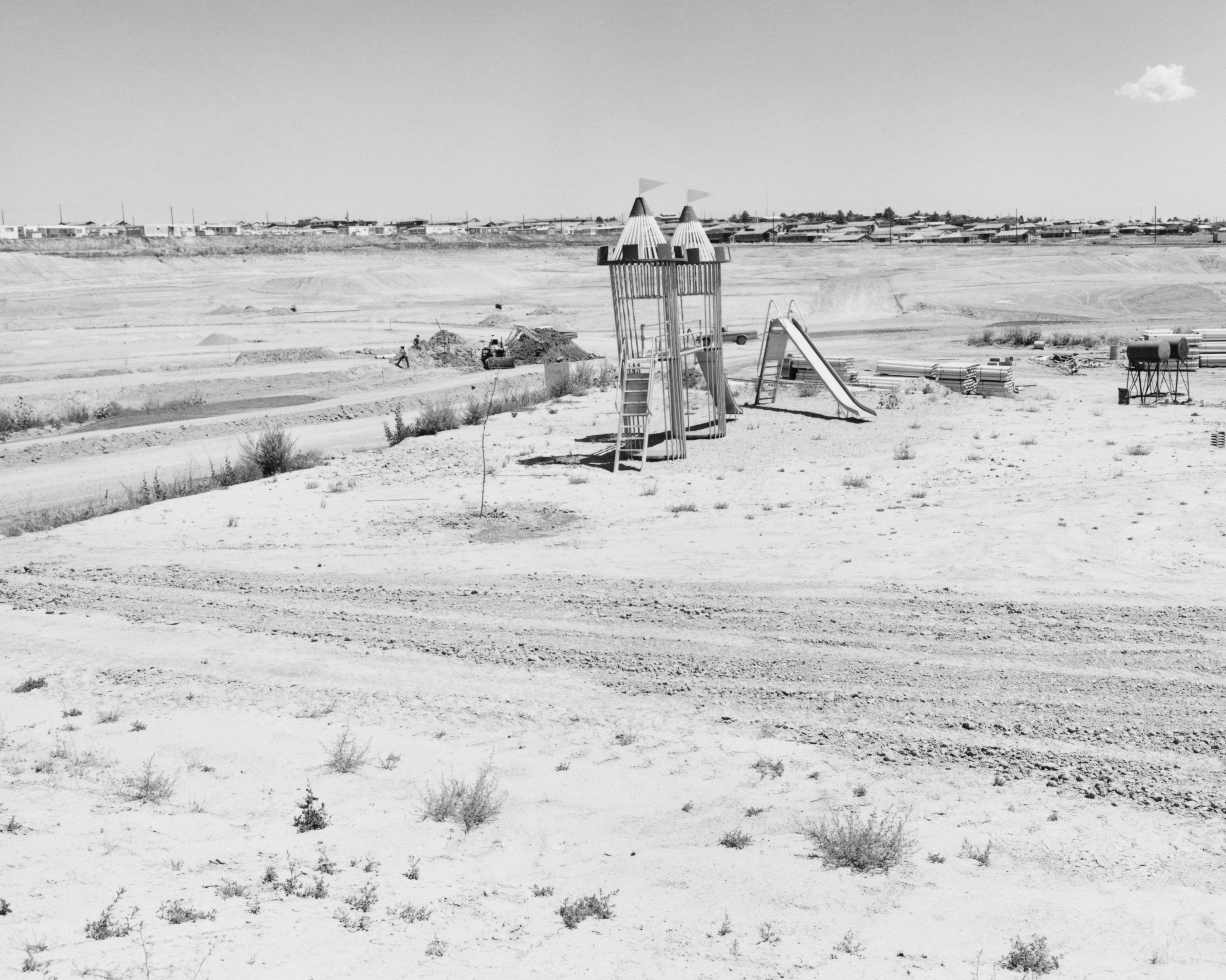 Black and white photograph of a developing playground within an empty dirt lot with three construction workers