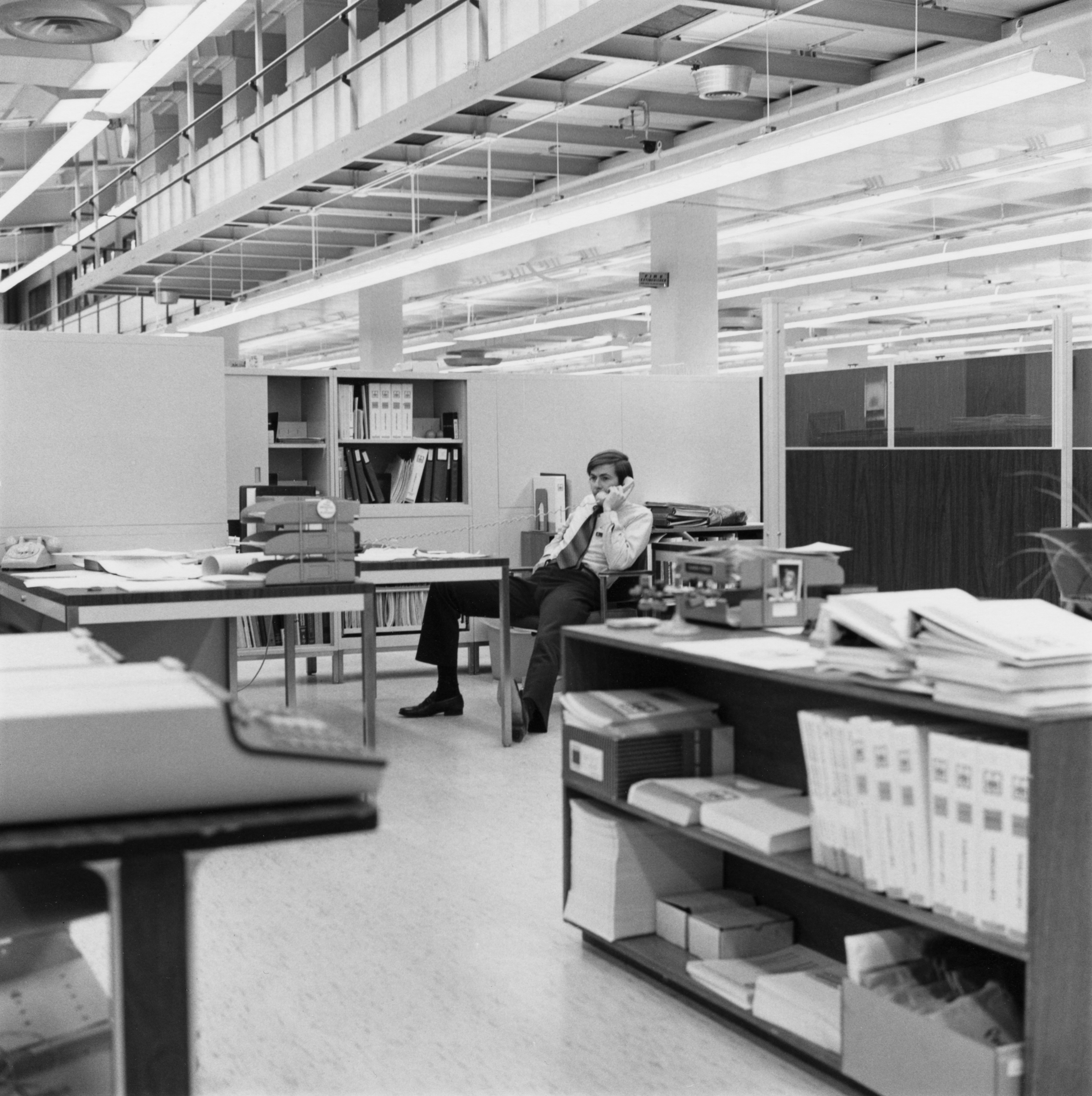 Black and white photograph of a seated figure on the phone behind a desk within an office