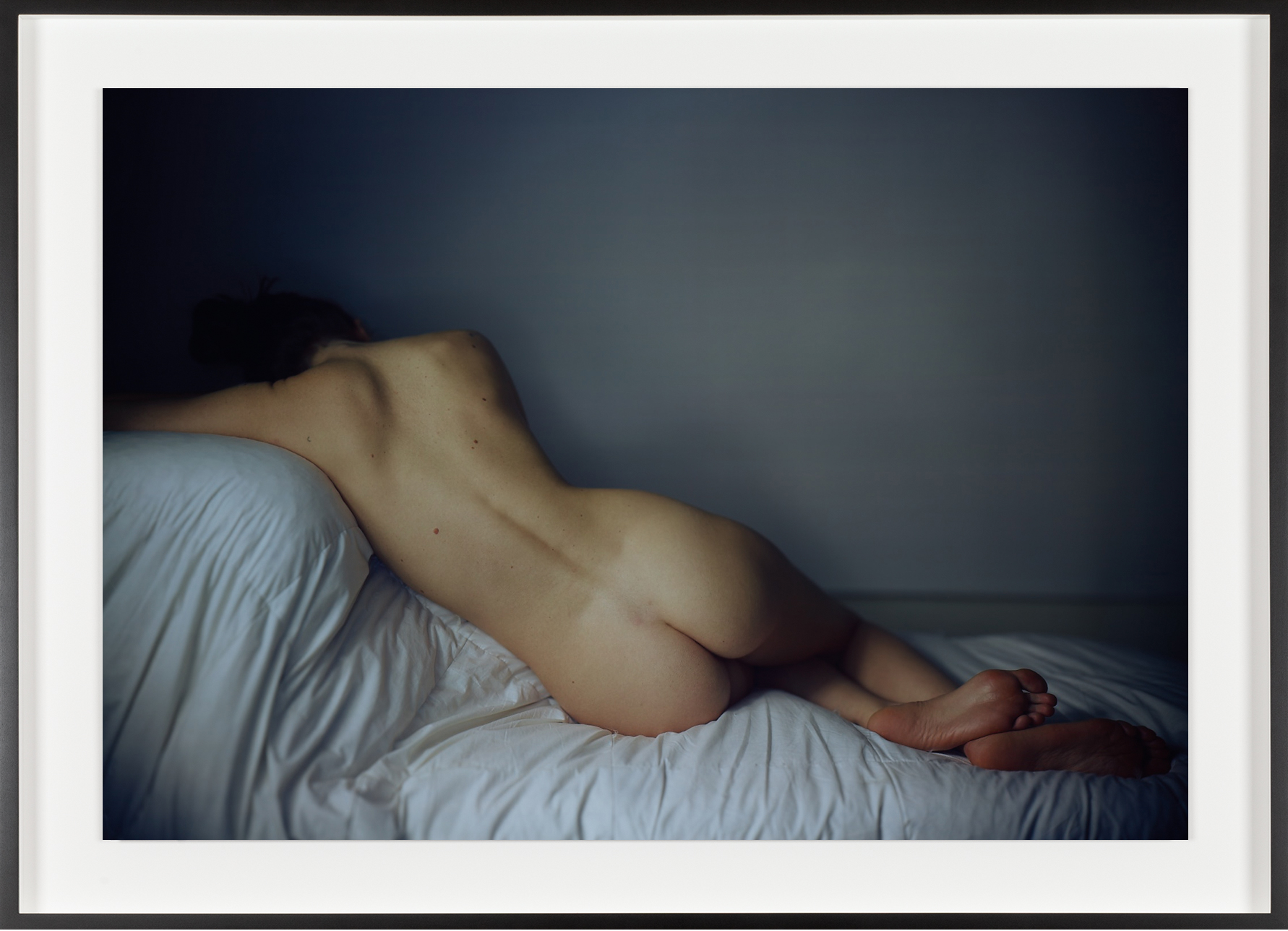 Color photograph of a nude figure from behind in repose on a blanketed white ottoman framed in black